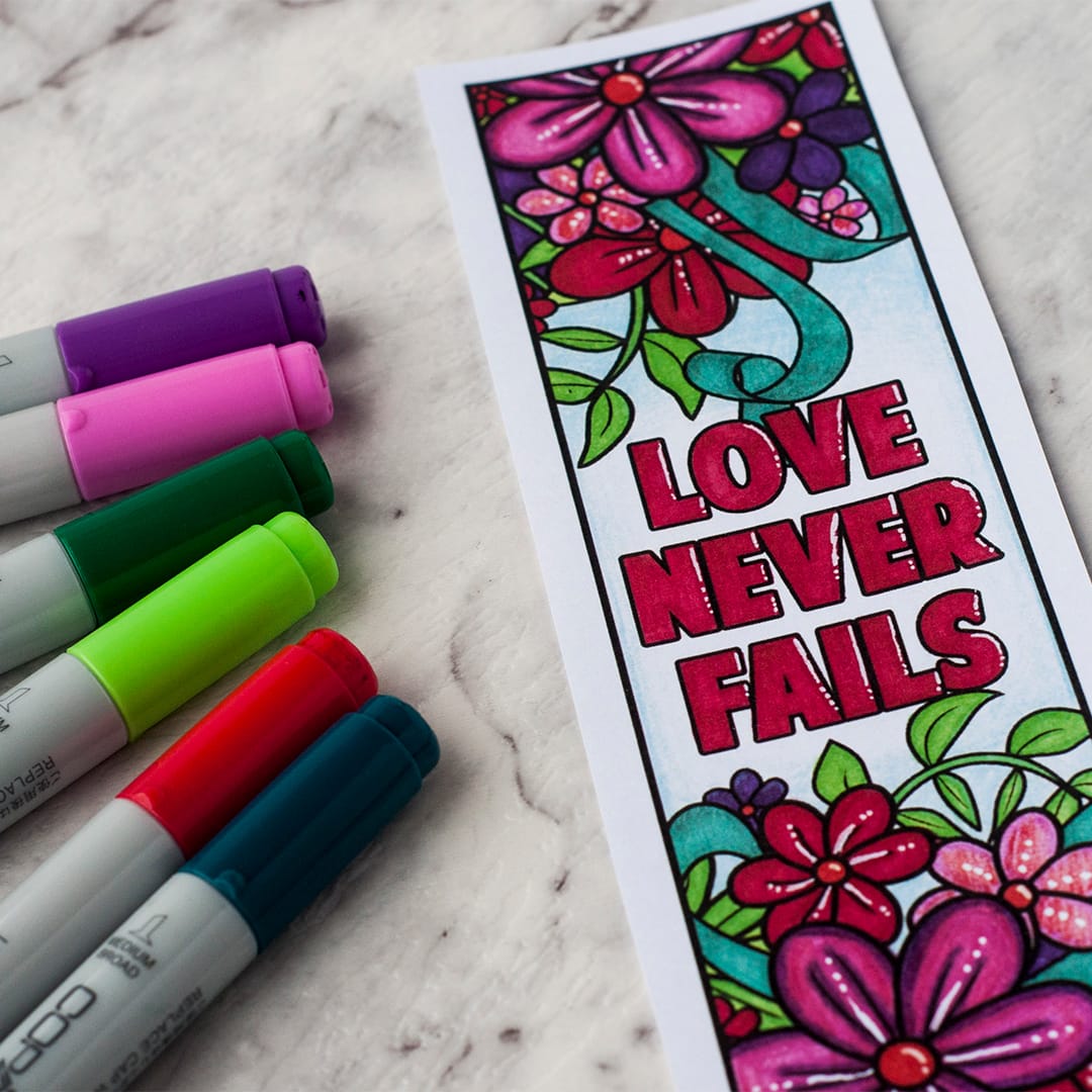 An image of a colored bookmark beside 6 copic markers. The bookmark reads 'Love Never Fails.'