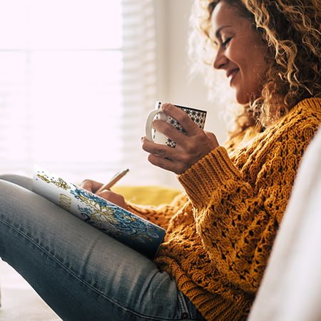 Happy cute lady at home write notes on a diary while drink a cup of tea and rest and relax taking a break. autumn colors and people enjoying home lifestyle writing messages or lists. Blonde curly beautiful lady sit down in the house